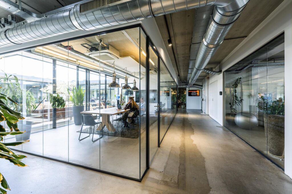 Modern office interior at Johan van Hasseltweg with glass meeting rooms, industrial ceilings, and indoor plants.